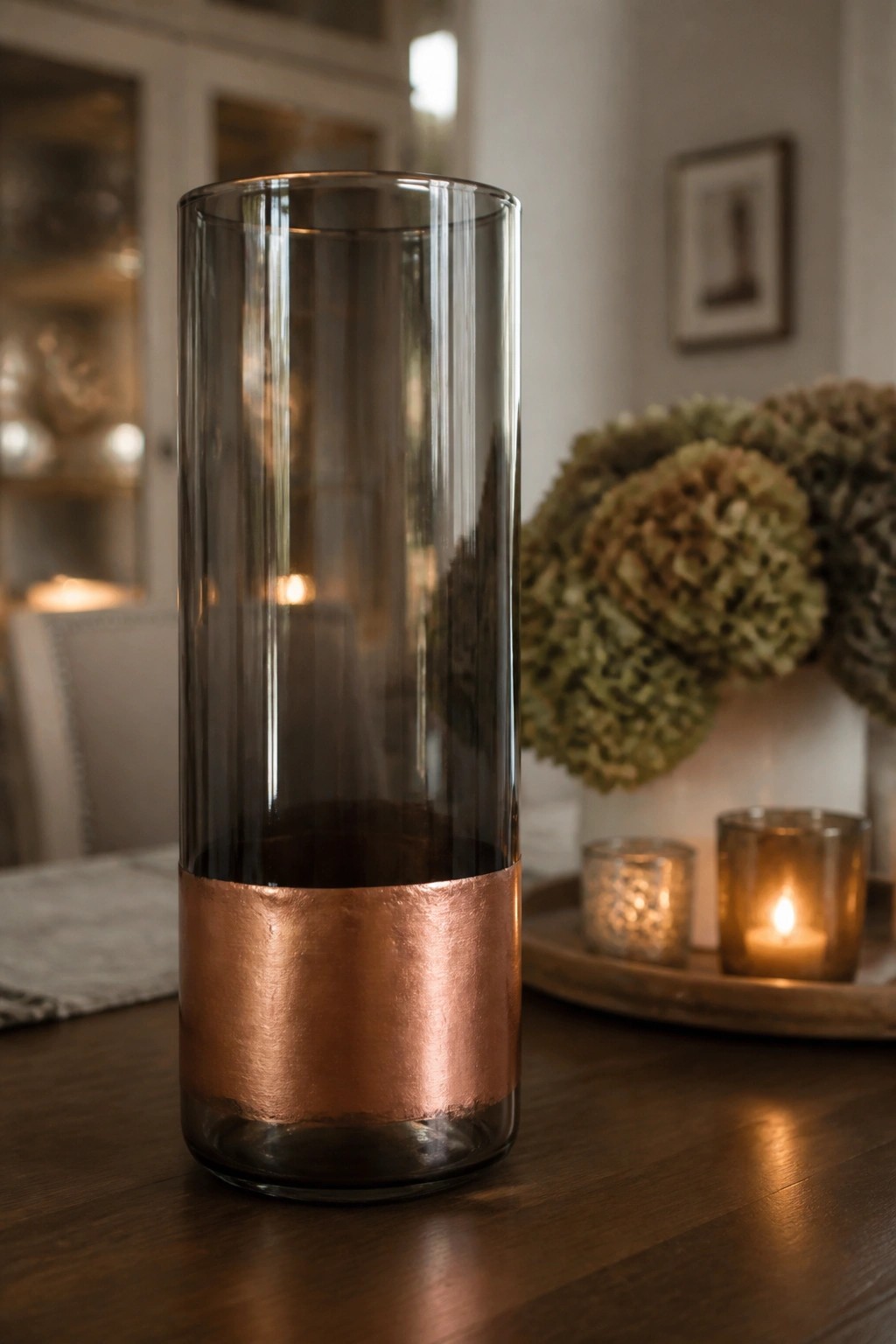 Tall glass vase with copper band on wooden table near lit candles and dried flowers.