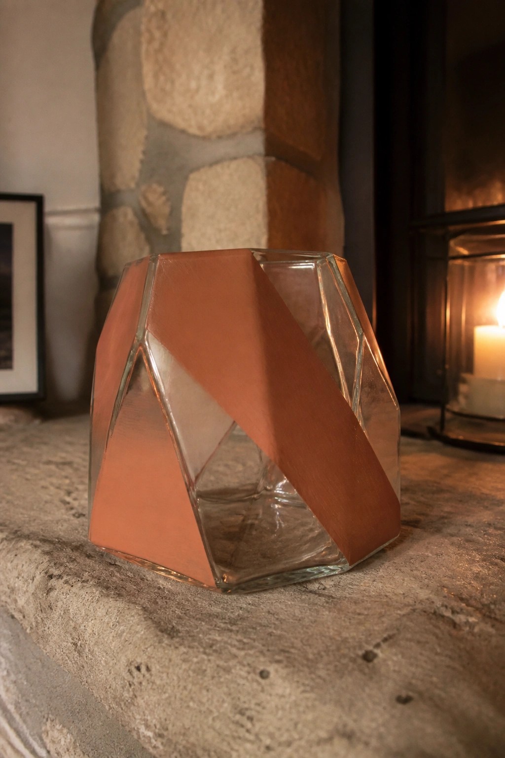 Faceted glass vase with copper panels on stone hearth beside lit candle