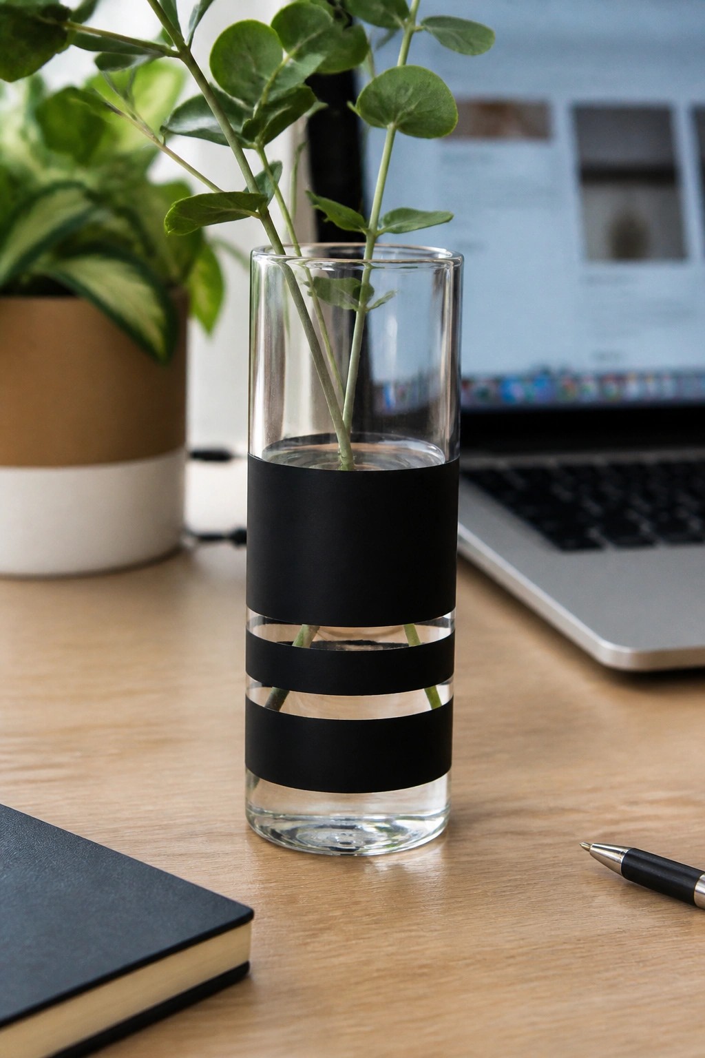 Clear glass vase with black stripes holding green plant stems on wooden desk