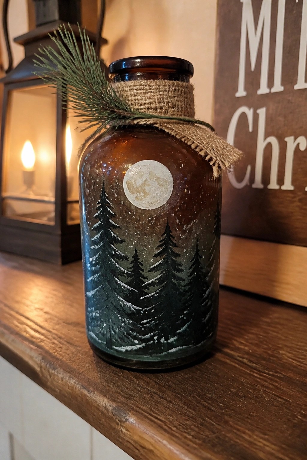 Amber glass bottle painted with snowy pines and moon, topped with burlap and greenery