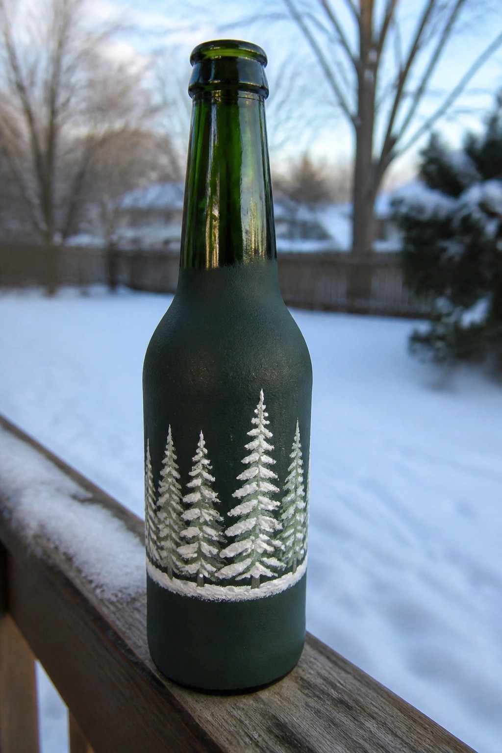 Green bottle painted with white pine trees on snowy wooden railing