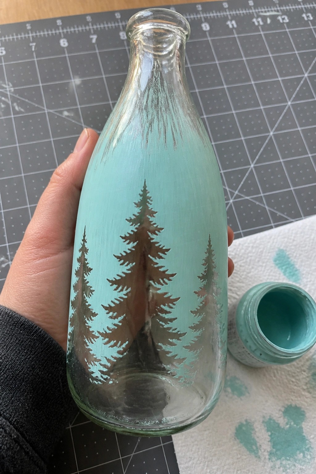 Hand holding light blue bottle with pine tree silhouettes beside open paint jar on mat.