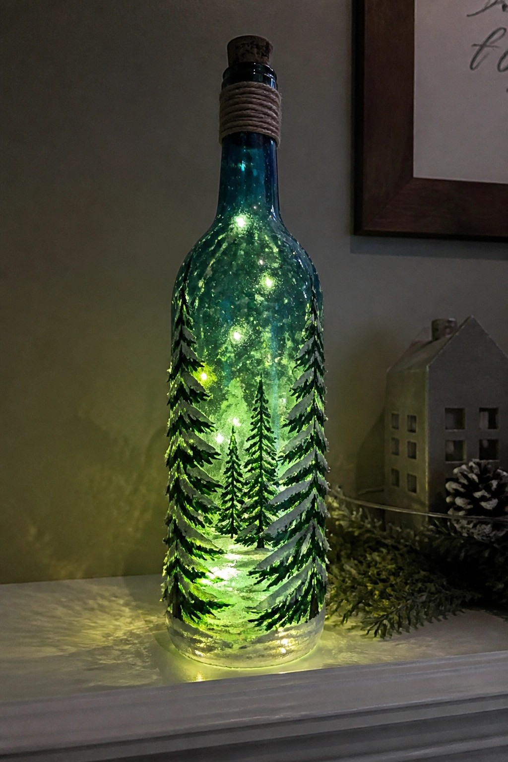 Glowing blue-green bottle with painted pine trees and internal lights on mantel.