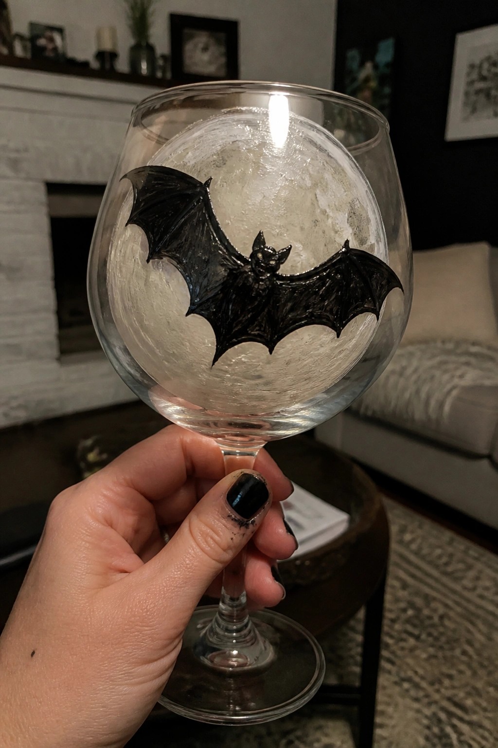 Hand with black nail polish holding wine glass with black bat on moon design.