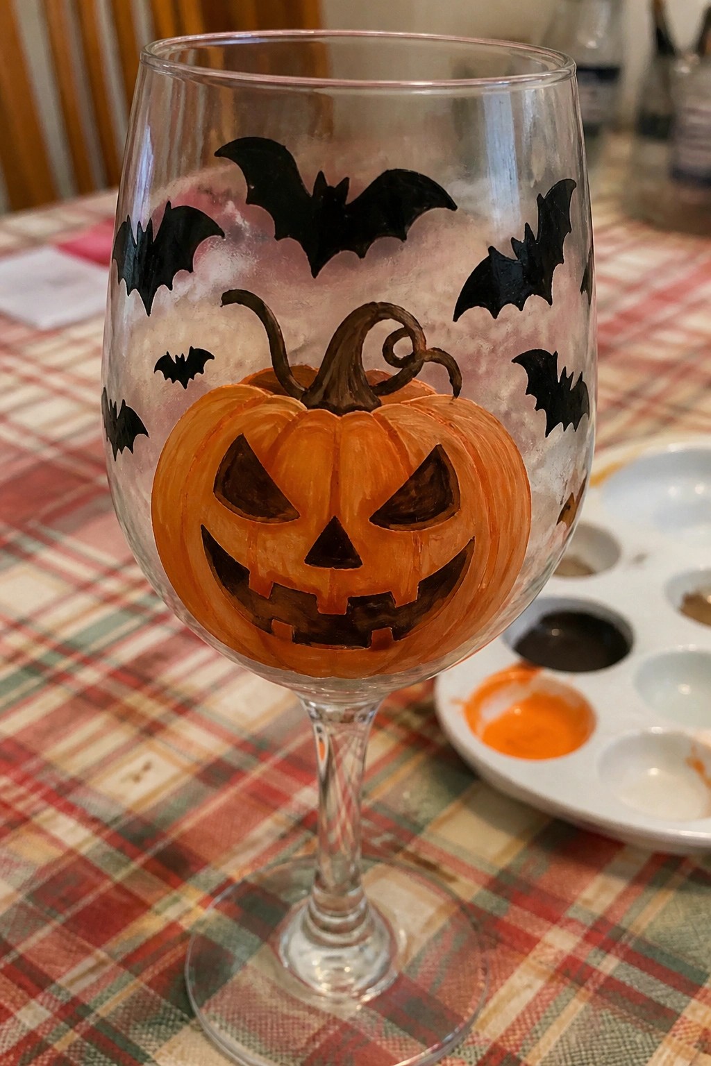 Painted wine glass with orange jack-o'-lantern, black bats on plaid tablecloth.