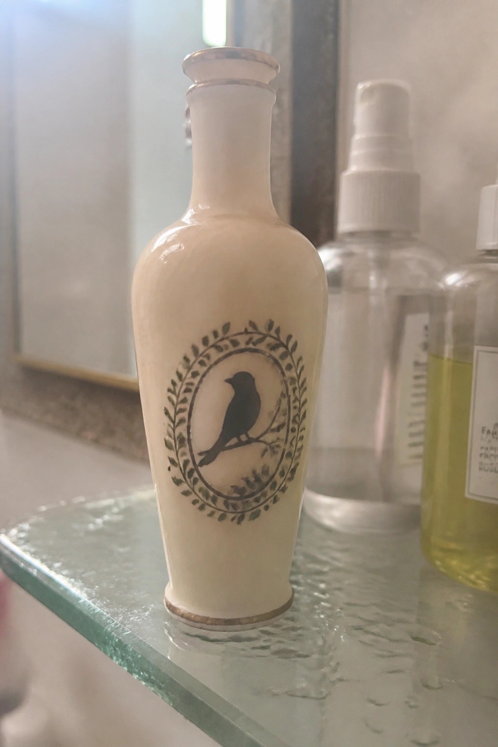 White ceramic bottle with black bird in leafy wreath and gold trim on glass shelf.