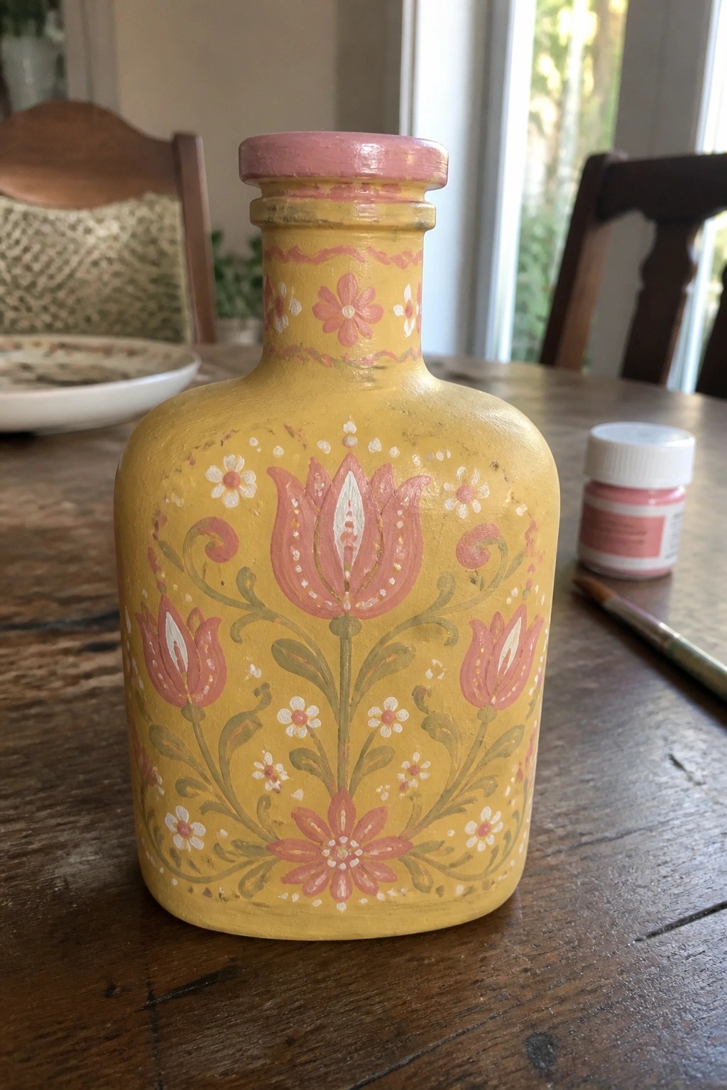 Yellow hand-painted bottle with pink tulips beside paint jar and brush on table