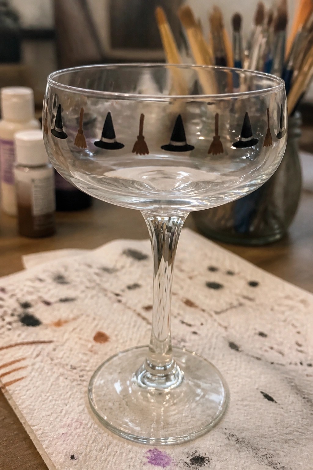Clear glass coupe painted with witch hats and brooms on paint-splattered paper towel.