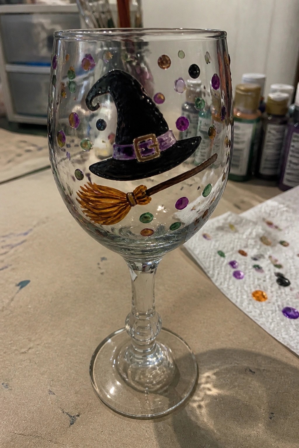 Wine glass painted with black witch hat, broomstick, and colorful scattered gems.