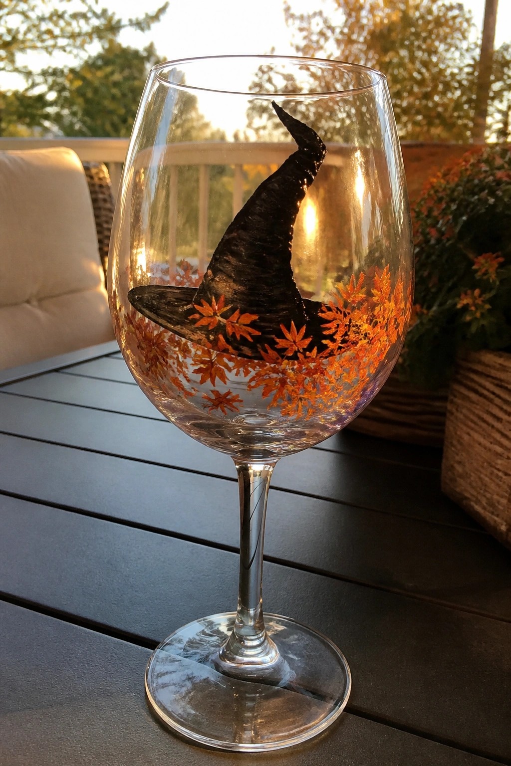 Wine glass with black witch hat and orange maple leaves on wooden table at sunset