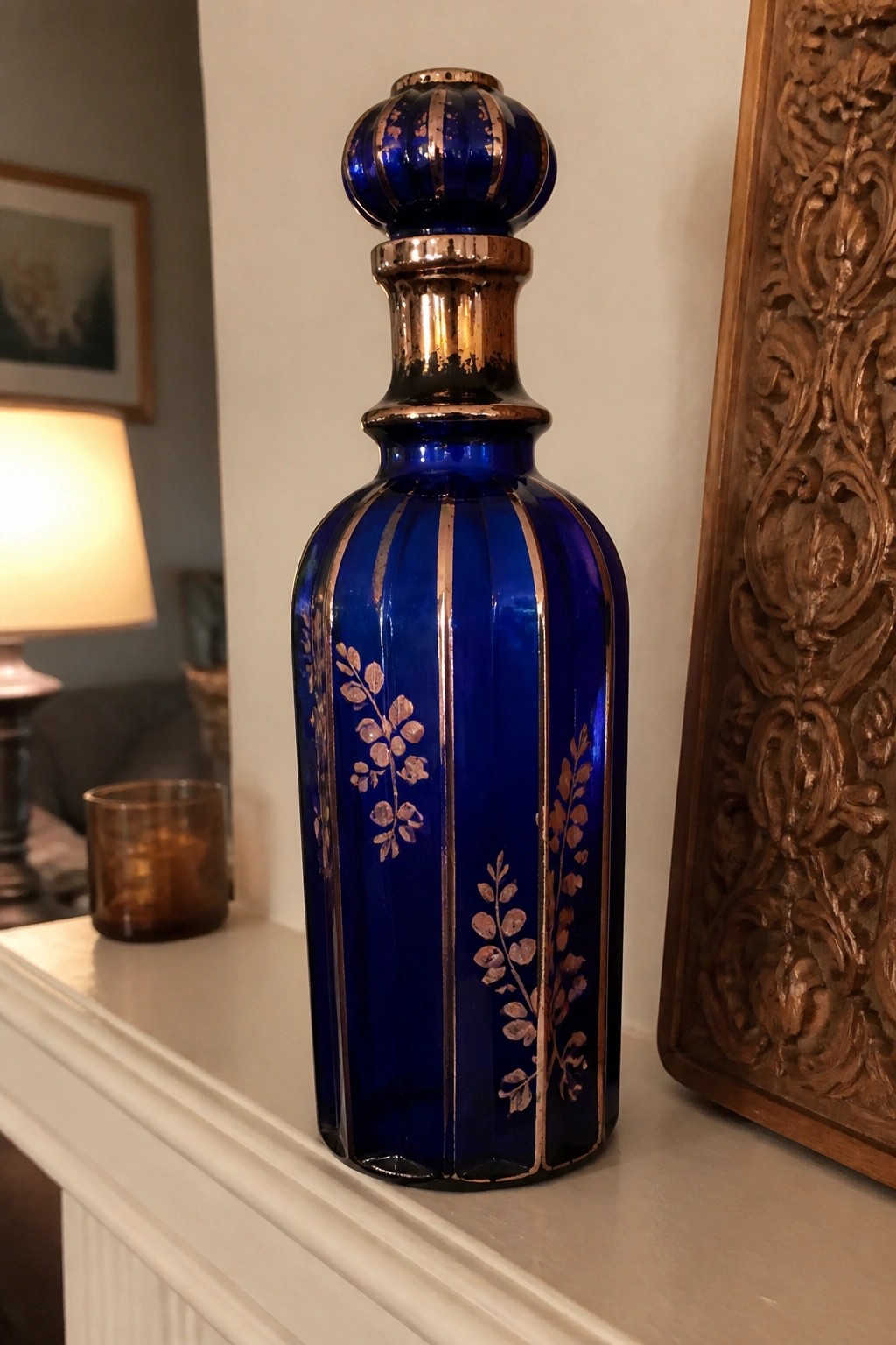 Deep blue glass bottle with gold stripes, floral motifs, and ornate stopper on mantel