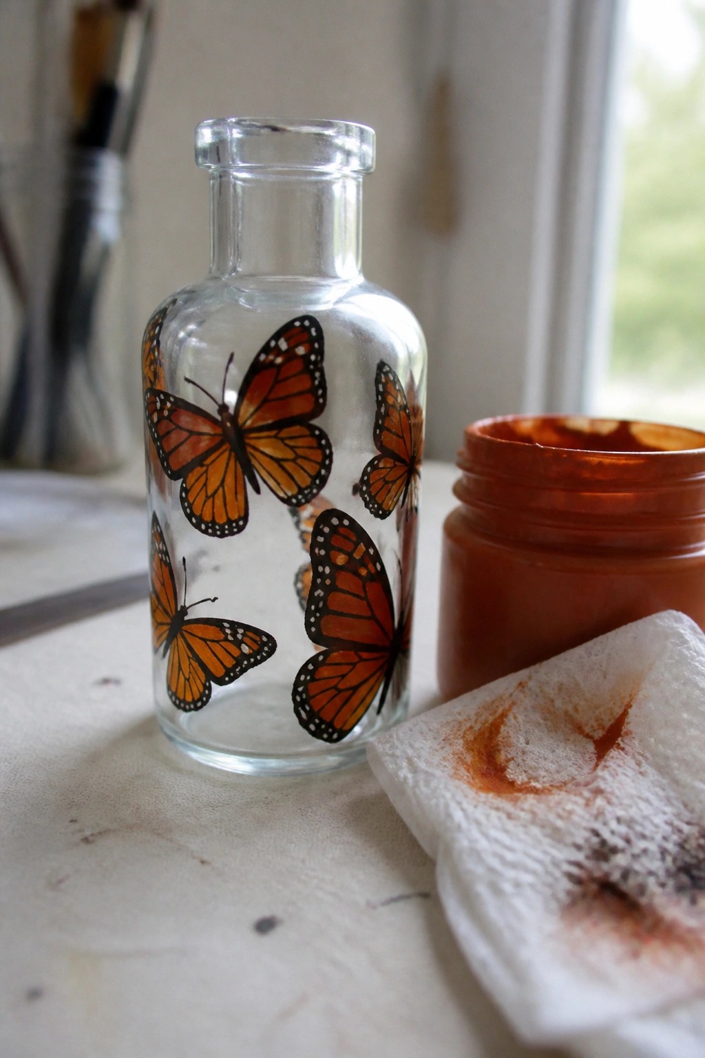 Glass bottle with orange monarch butterflies beside open paint jar and stained cloth