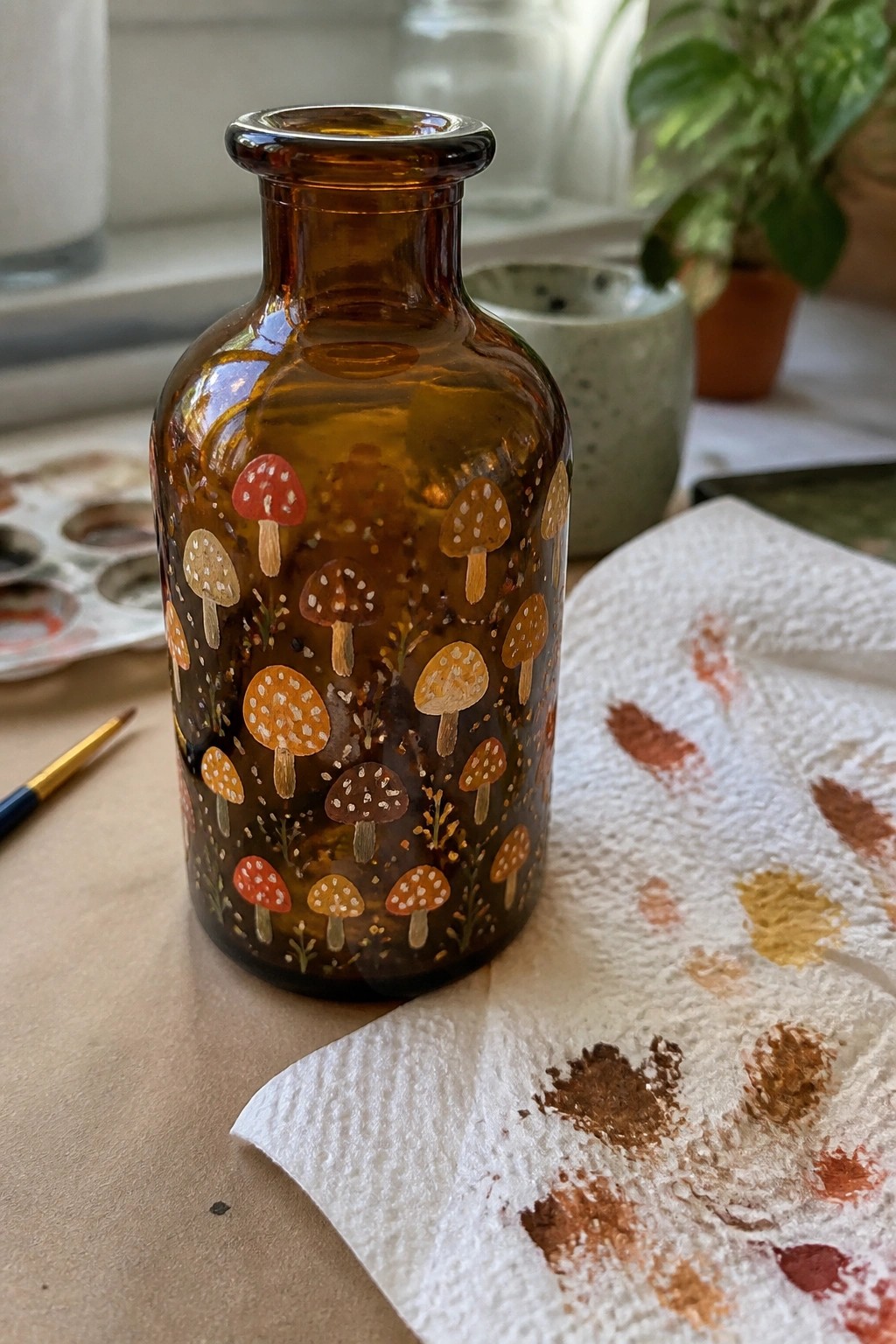 A brown glass bottle with painted mushrooms in red, orange, and brown sits beside a paint palette and stained paper towel.