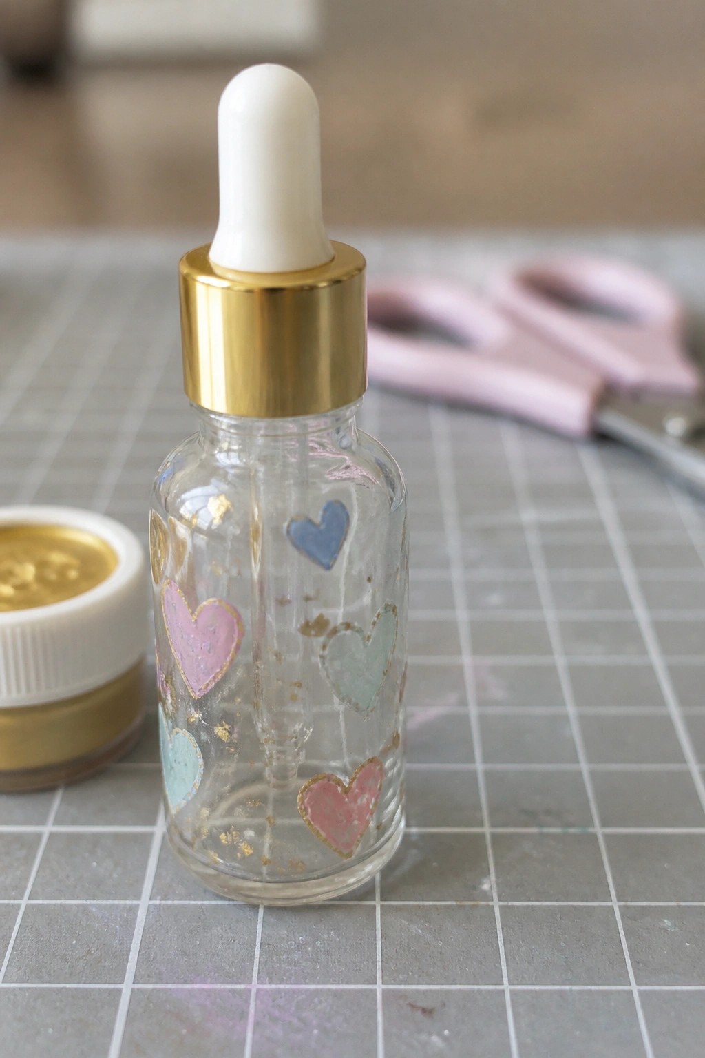 Clear dropper bottle with pastel hearts and gold foil beside paint jar and scissors