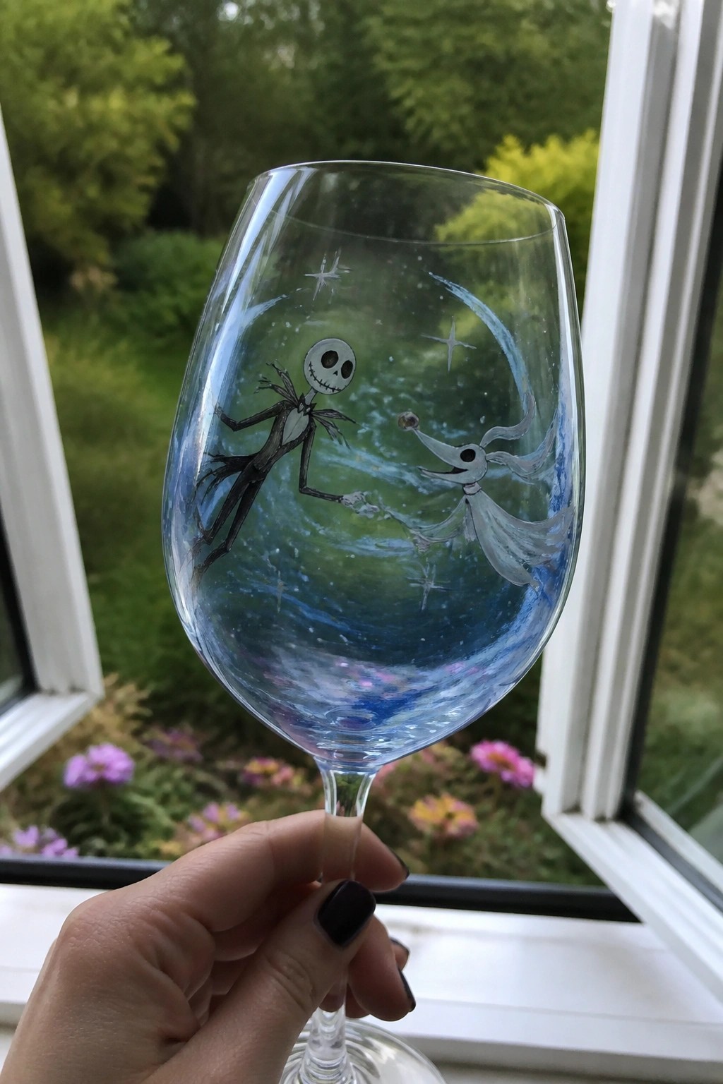 A hand holds a clear wine glass painted with Jack Skellington and Sally against a blue starry background.