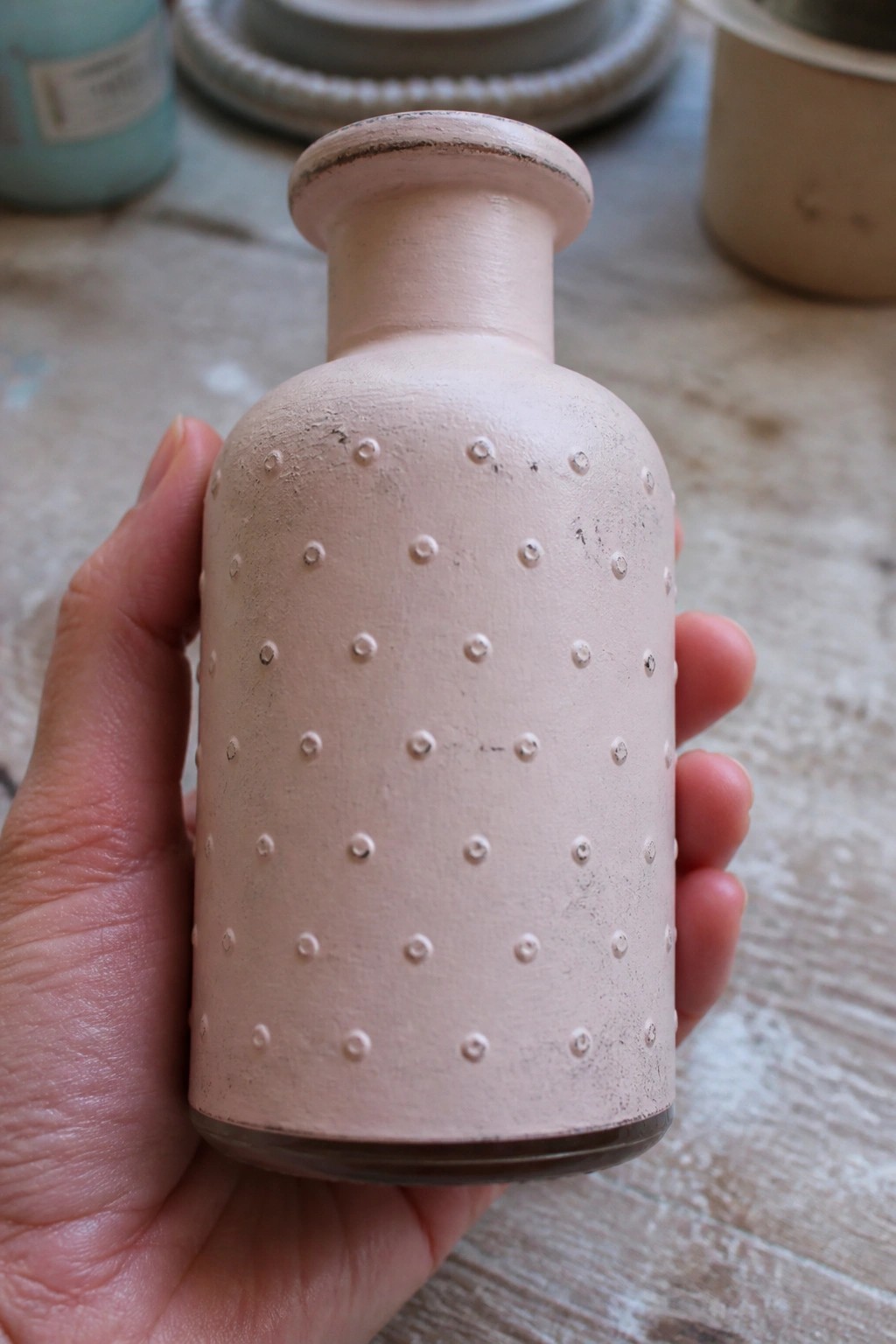 Hand holding pale pink ceramic bottle with raised dotted pattern on wood surface