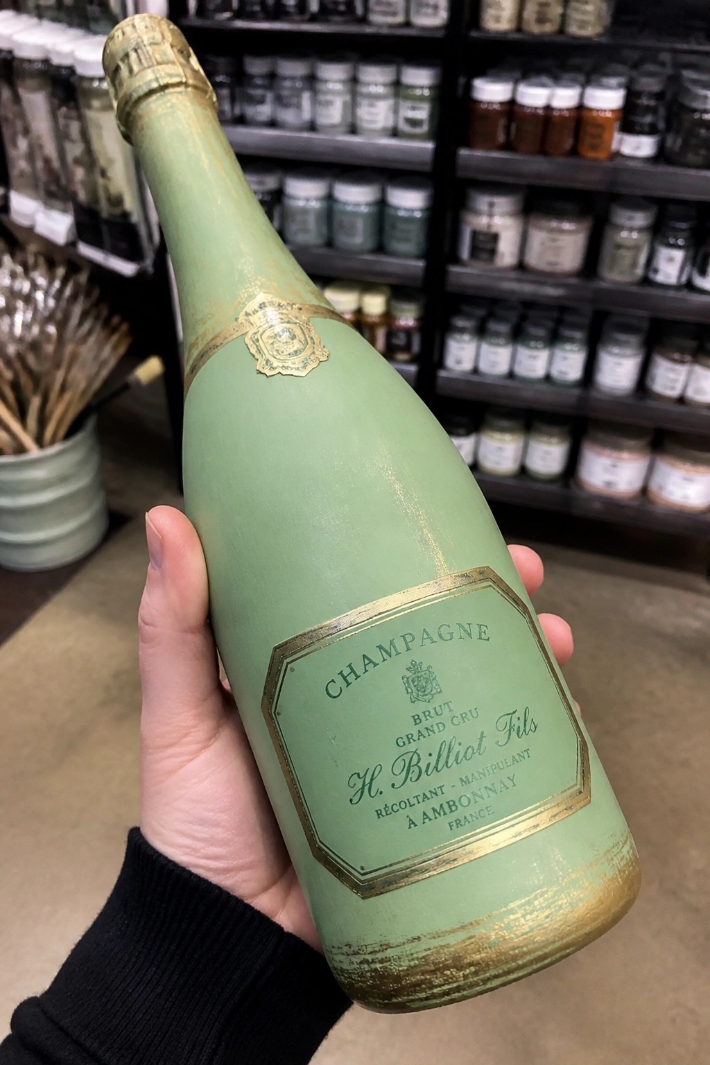 Hand in black sleeve holding pale green champagne bottle with gold label in store