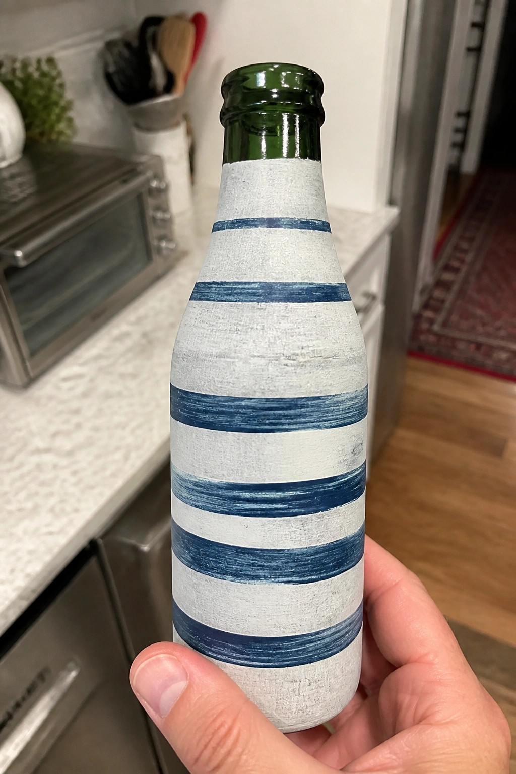 Hand holding green glass bottle painted white with blue horizontal stripes