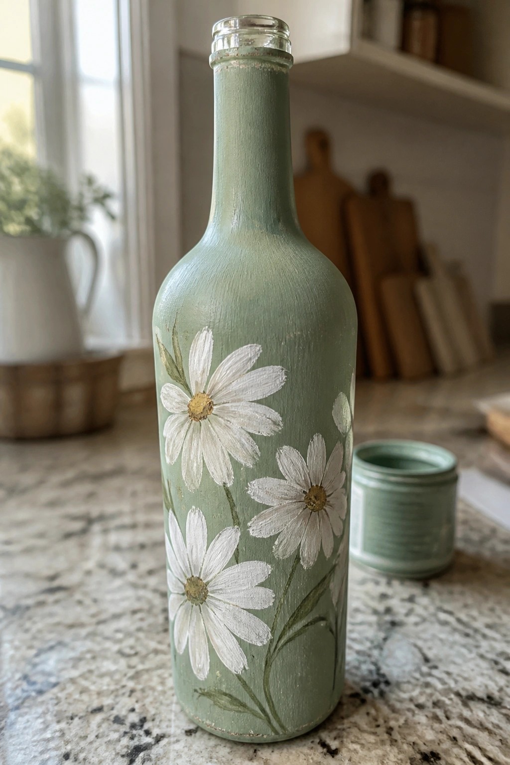 Sage green painted bottle with white daisies beside open paint jar on counter