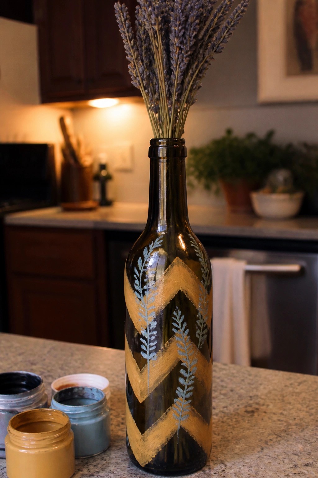 Painted dark bottle with gold chevrons and blue leaves holding lavender beside paint jars