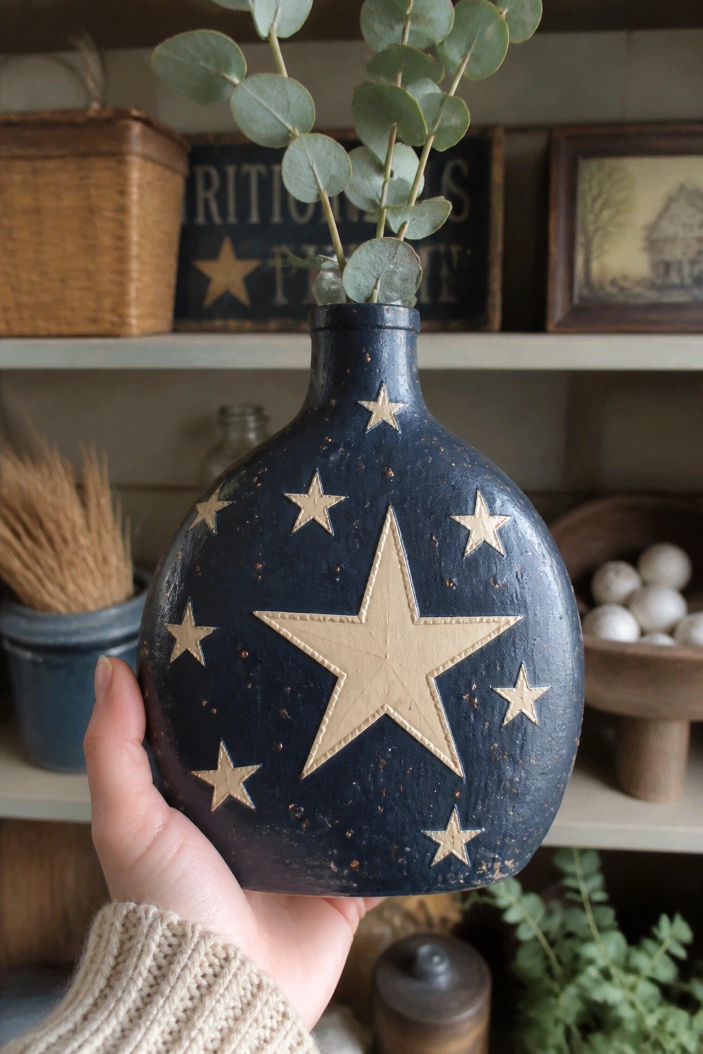 Hand in beige sweater holds navy blue vase with gold stars and eucalyptus.