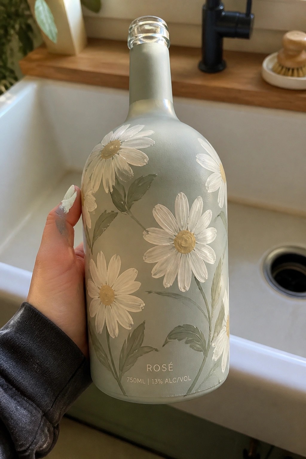 Hand holding sage-green bottle with white daisies above kitchen sink