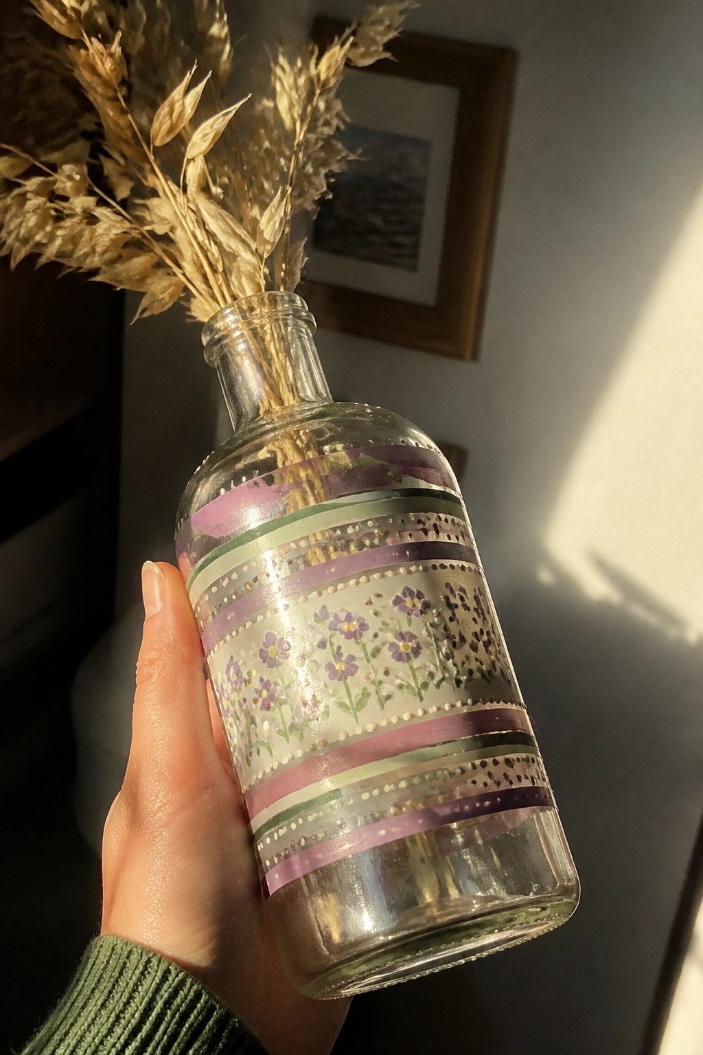 Hand in green sweater holding floral washi-taped glass bottle with dried grasses