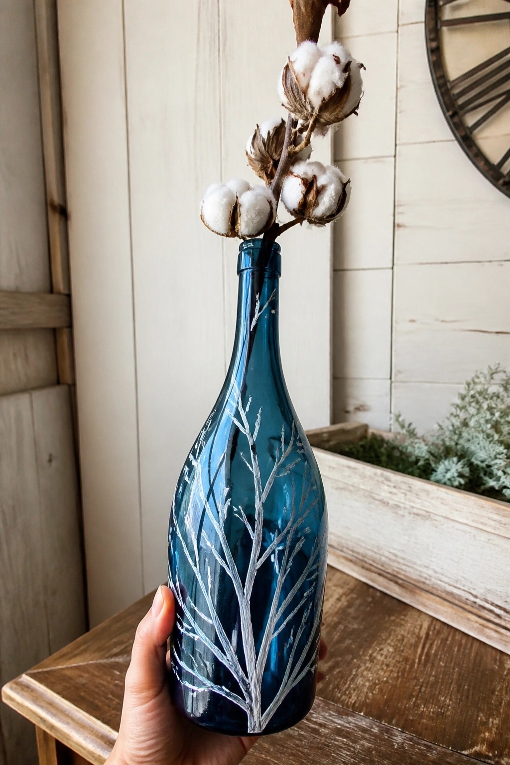 Hand holding teal glass bottle with white tree design and cotton bolls