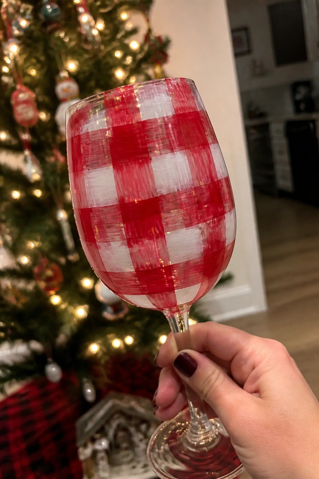 Hand holding red-and-white checkered wine glass before a lit Christmas tree.