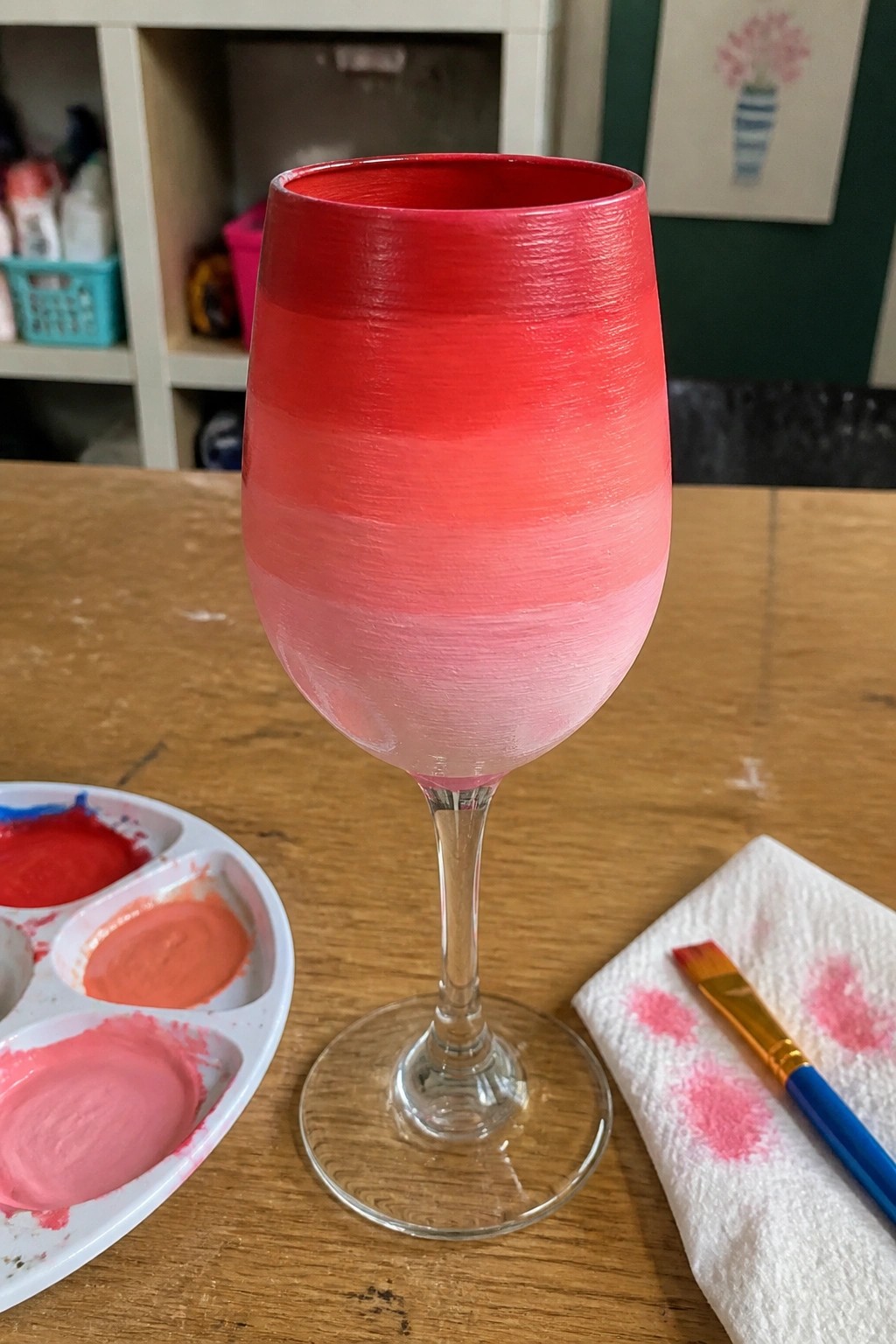 Ombre red-to-pink painted wine glass on wooden table with paint palette and brush