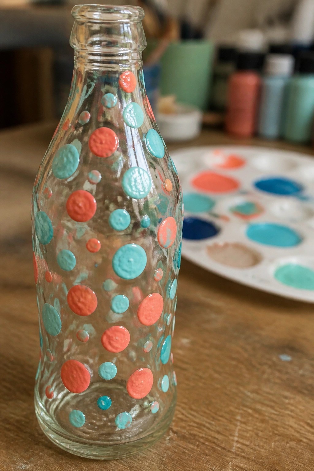 Glass bottle dotted with coral and turquoise paint beside colorful palette on wood table