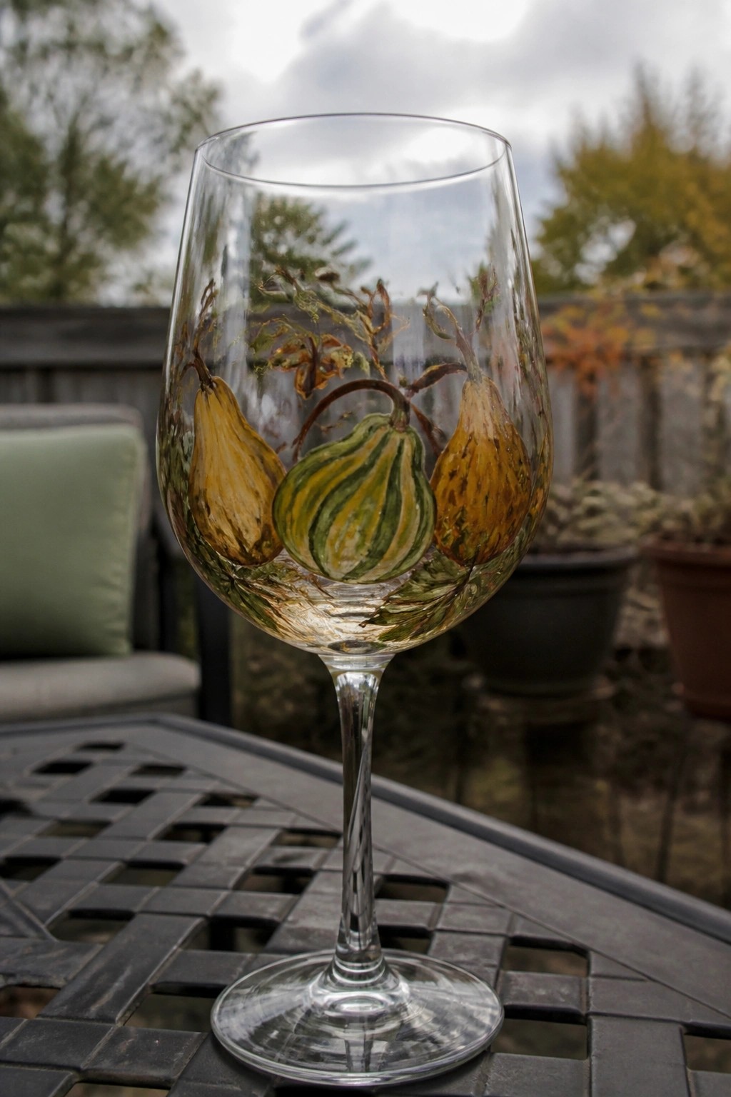 Hand-painted wine glass with yellow and green gourds on outdoor metal table.
