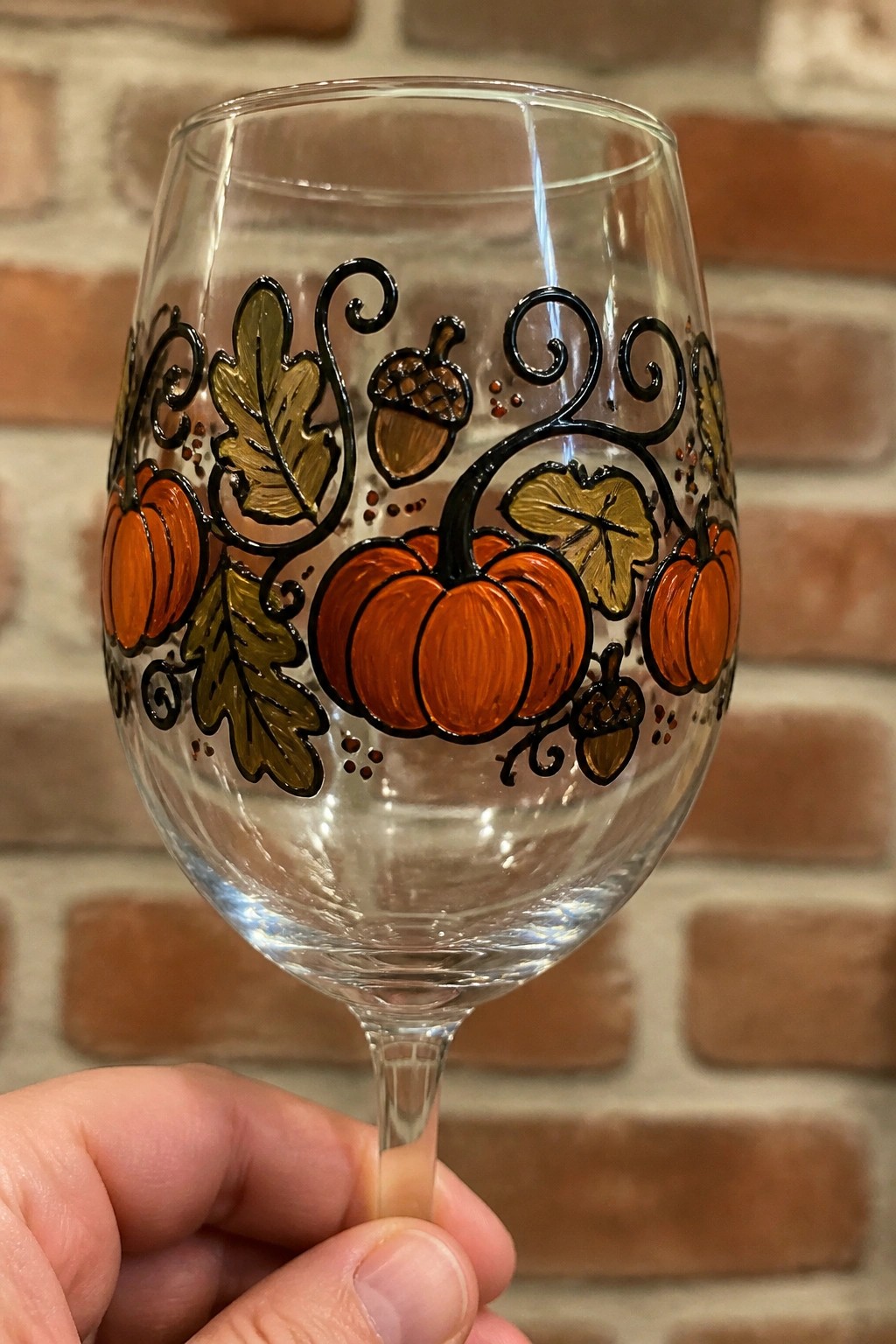 Hand holding wine glass painted with orange pumpkins, acorns, and autumn leaves.