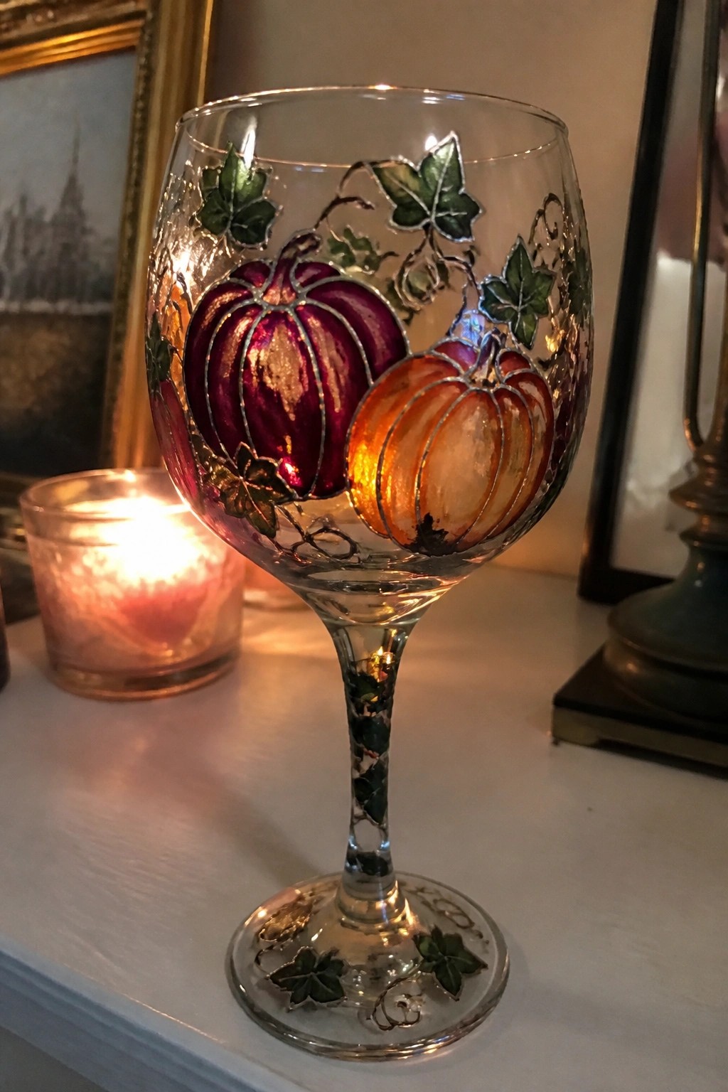 Ornate wine glass painted with purple and orange pumpkins and green ivy beside lit candle