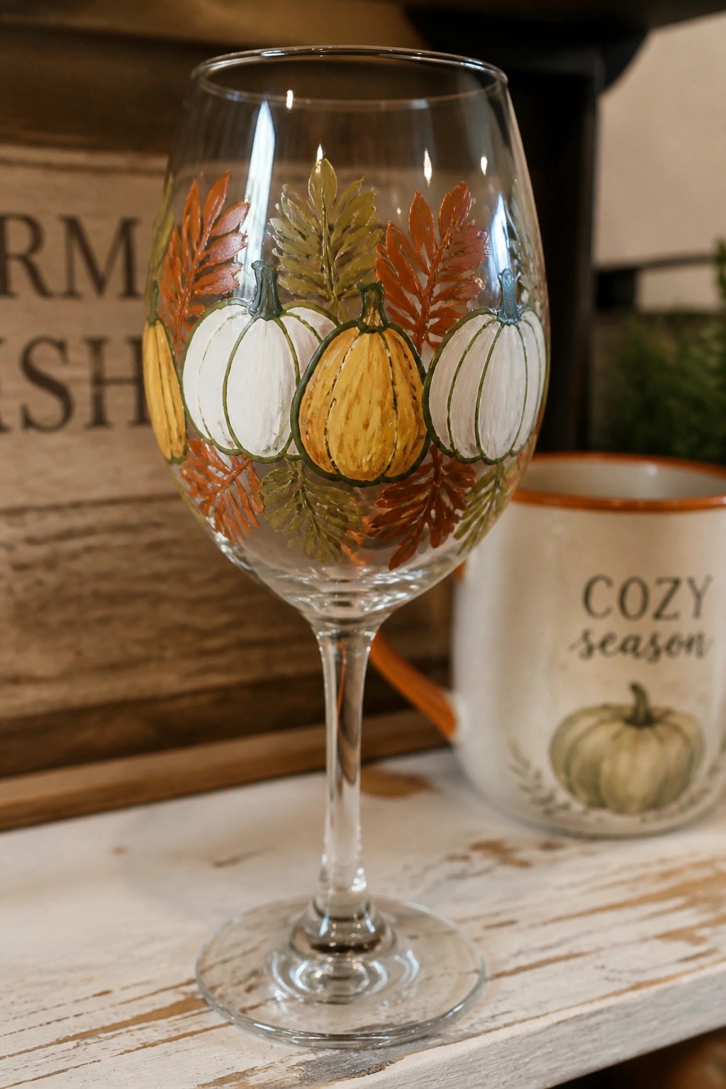 Painted wine glass with white and orange pumpkins amid autumn leaves.