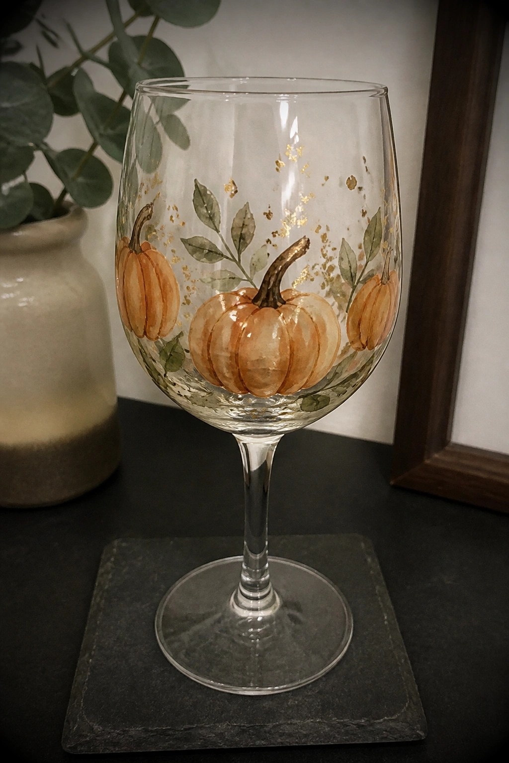 Decorated wine glass with pumpkins, leaves, and gold accents on dark coaster.