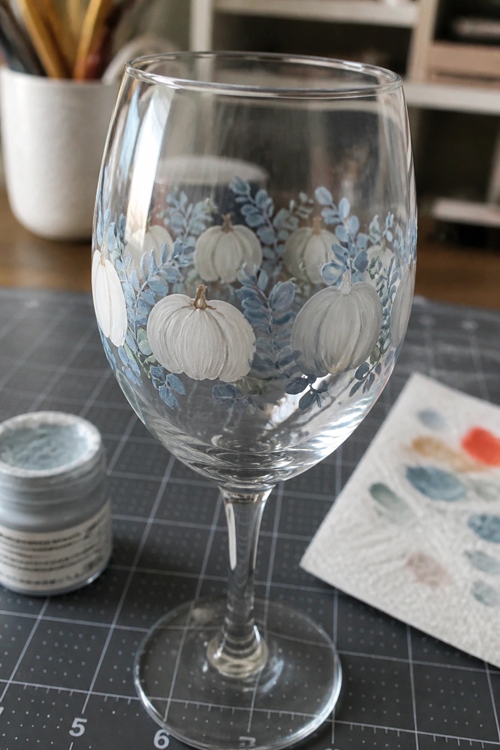Wine glass painted with white pumpkins and blue leaves on gridded mat