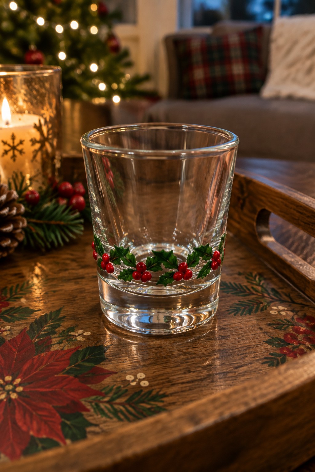 Holly-decorated glass on wooden tray beside lit candle and Christmas tree.