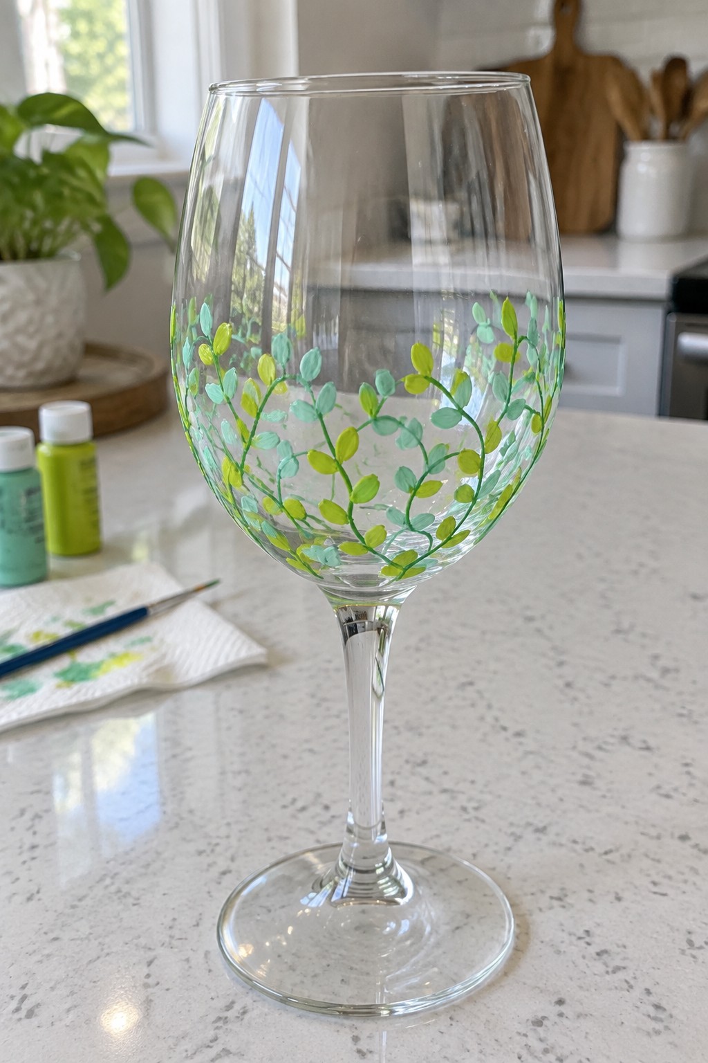 Hand-painted wine glass with green-yellow leafy vines on marble counter beside paints