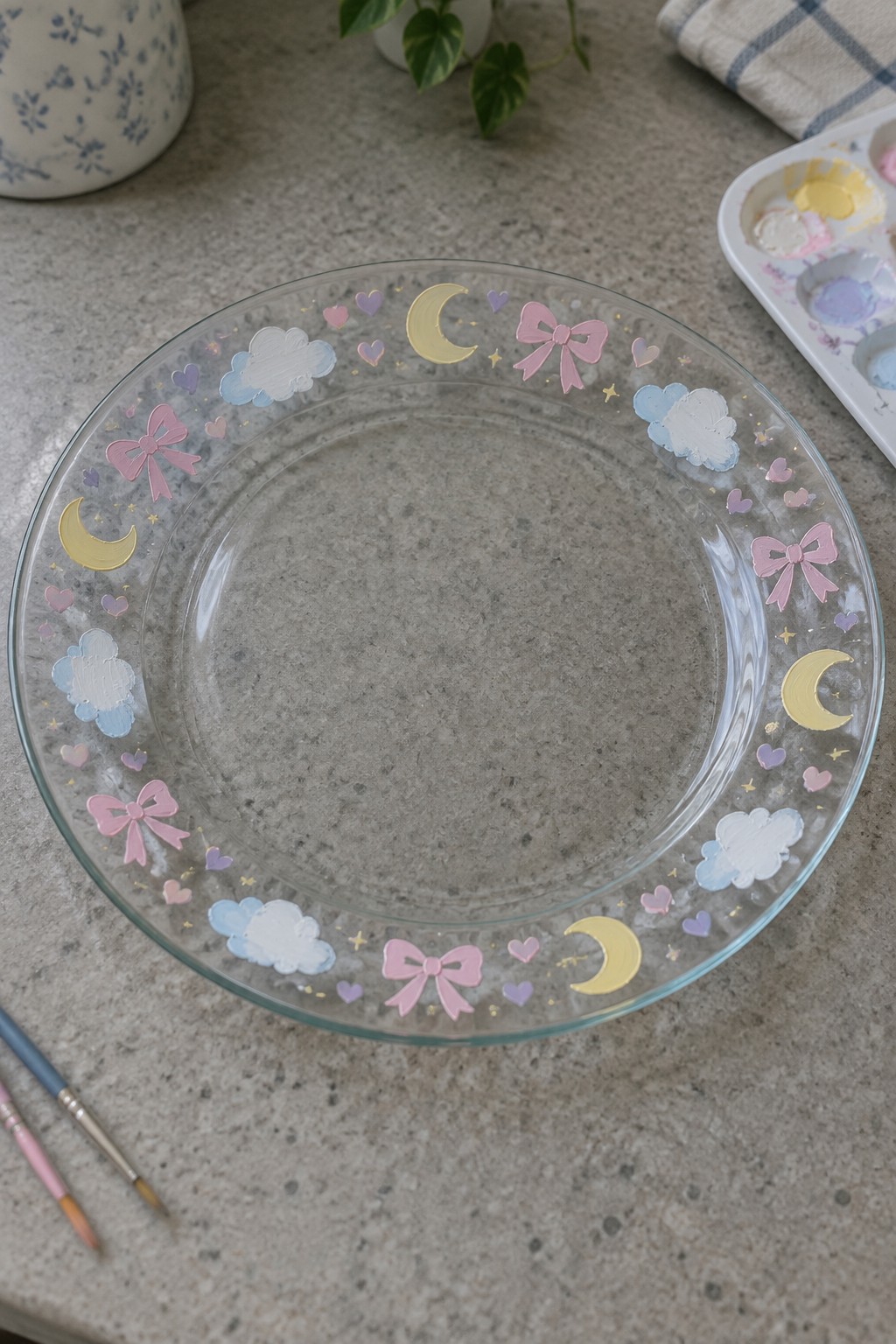 A clear glass plate painted with a border of pink bows, clouds, moons, hearts, and stars in pastel colors next to paint supplies.