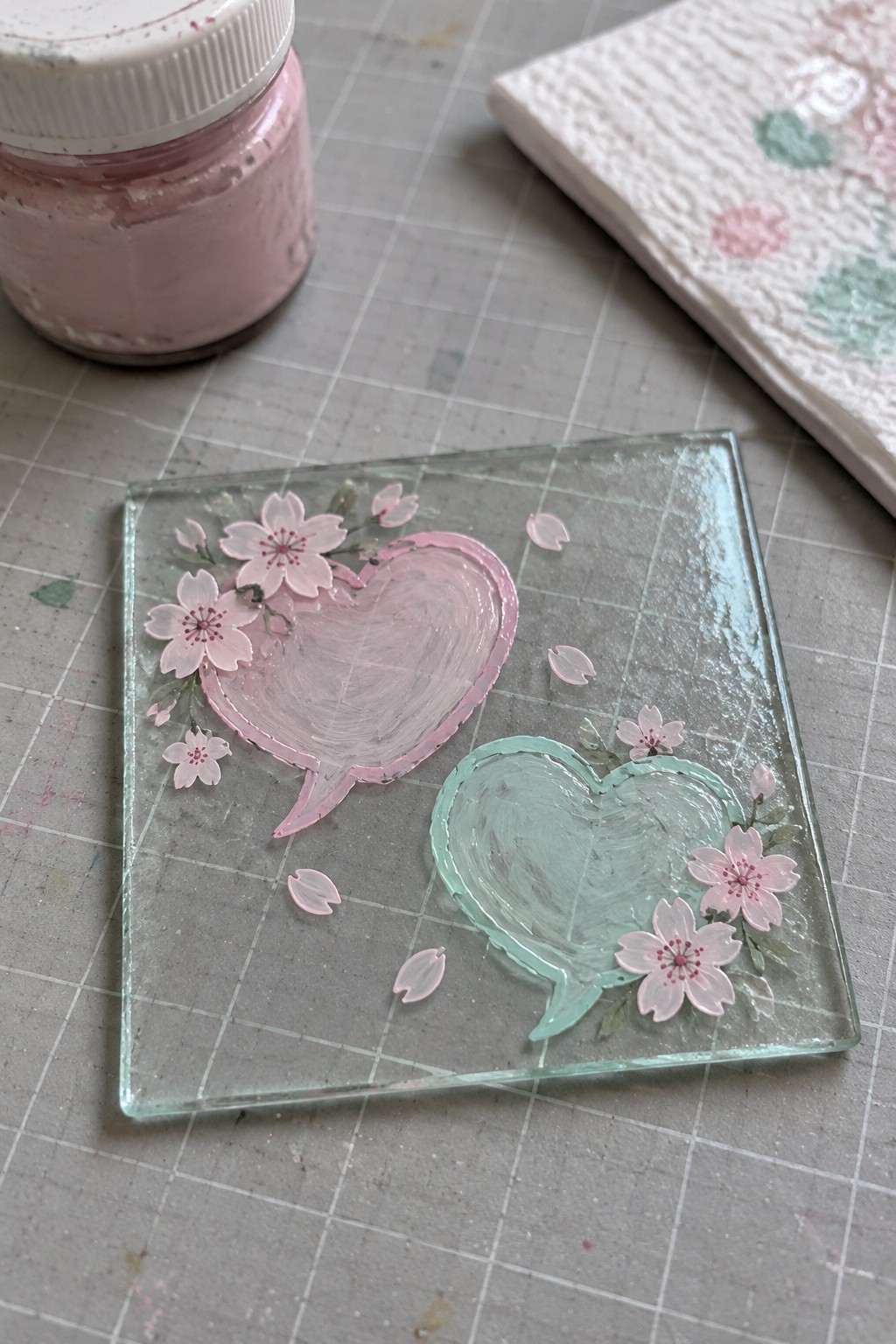 A square clear glass panel with a pink heart speech bubble and a mint green heart speech bubble, both surrounded by painted cherry blossoms and petals, sitting on a gridded mat beside a jar of pink paint.