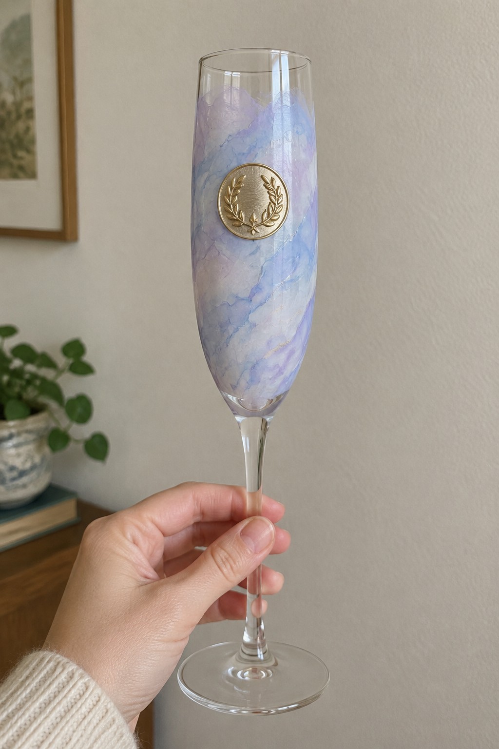 Hand in cream sweater holding purple-blue marbled champagne flute with gold laurel emblem.