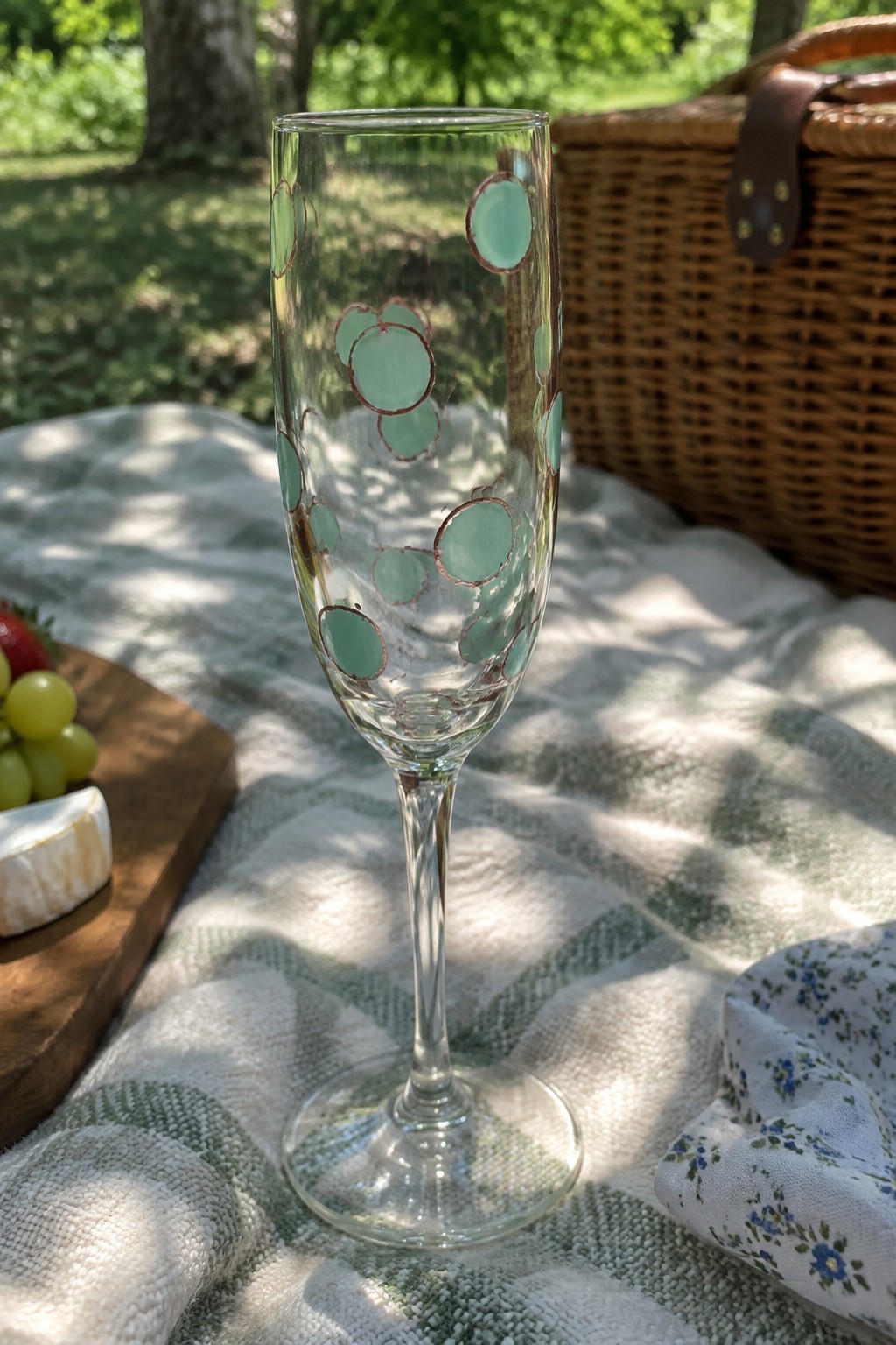Champagne flute with mint green circles on checkered picnic blanket near basket