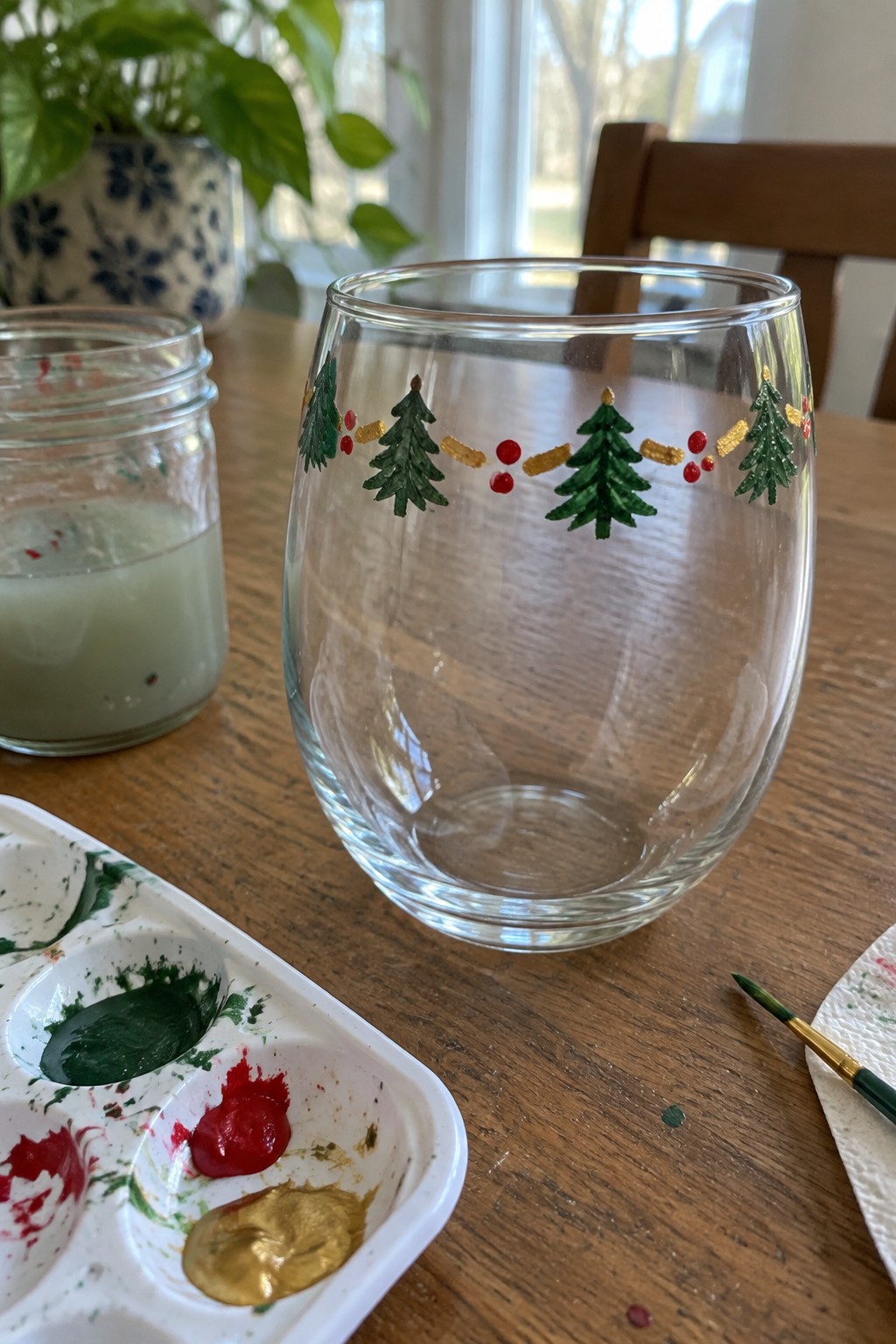 Stemless glass with painted Christmas trees, red dots, gold garland near paint palette