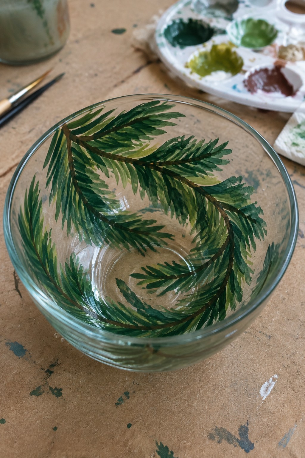 A clear glass bowl with green evergreen branches painted on it sits on a wooden table next to paint supplies.