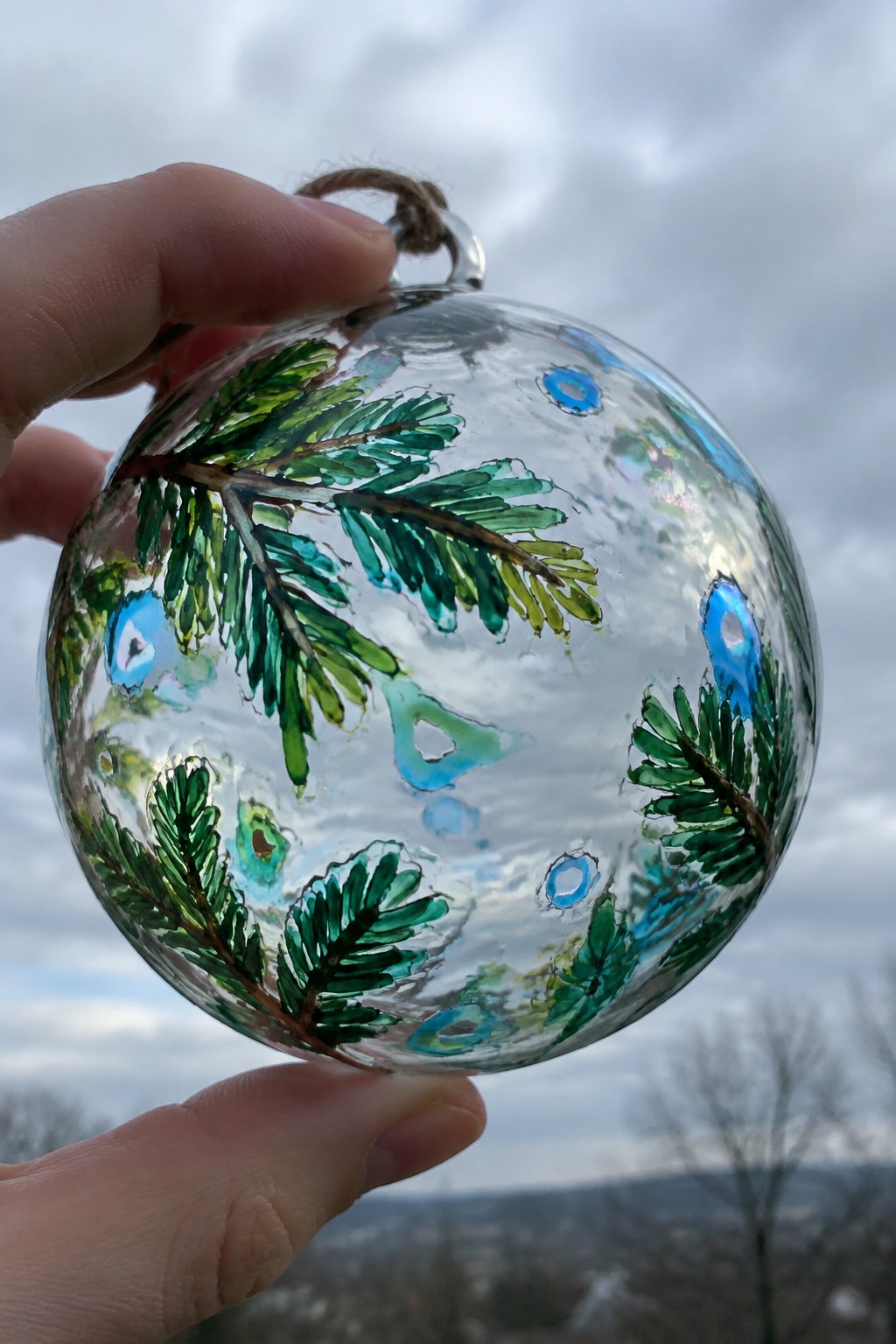 Hand holding clear glass ornament painted with green pine branches and blue accents