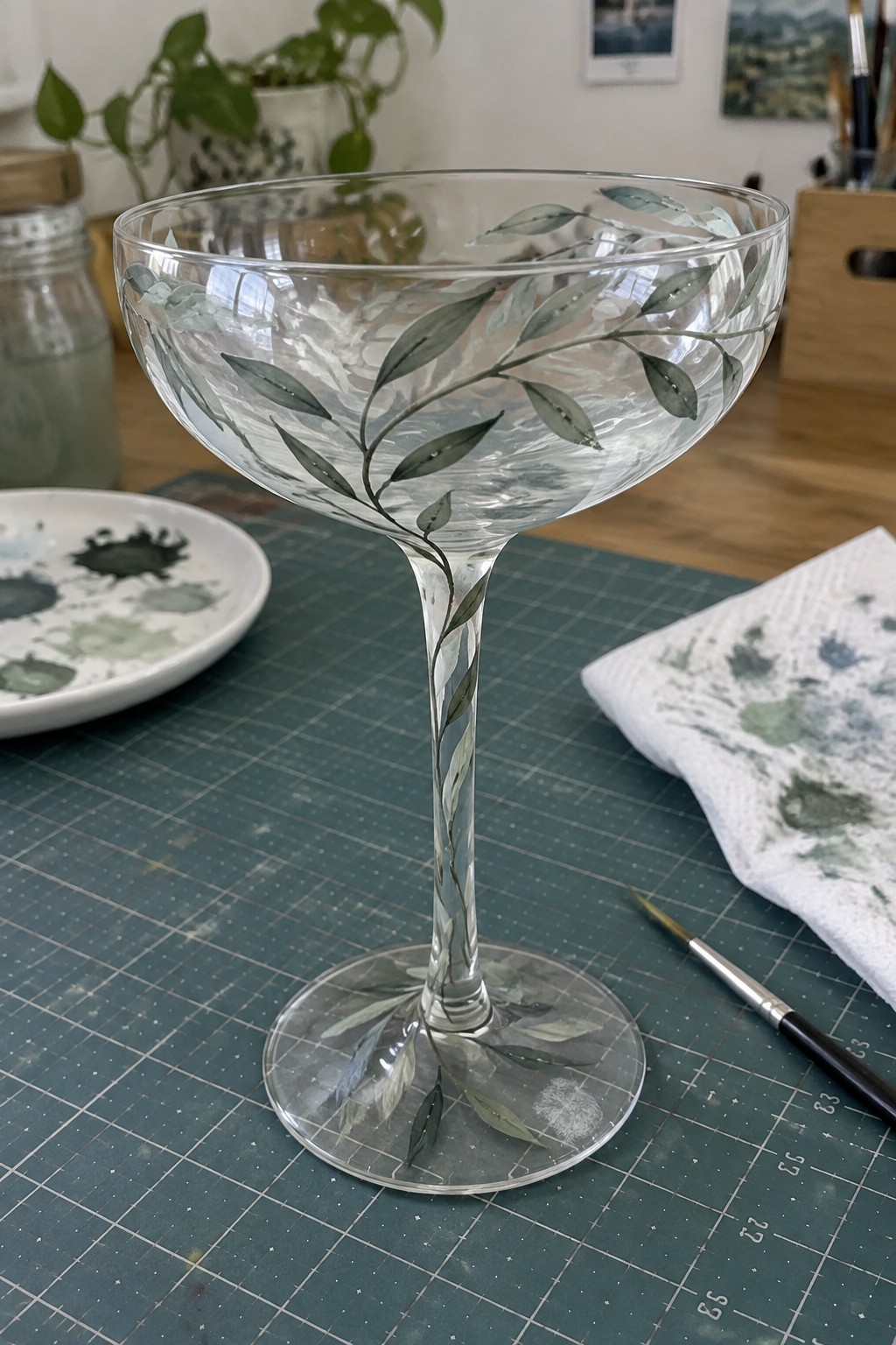 Glass coupe with green leaf painting on stem atop green cutting mat