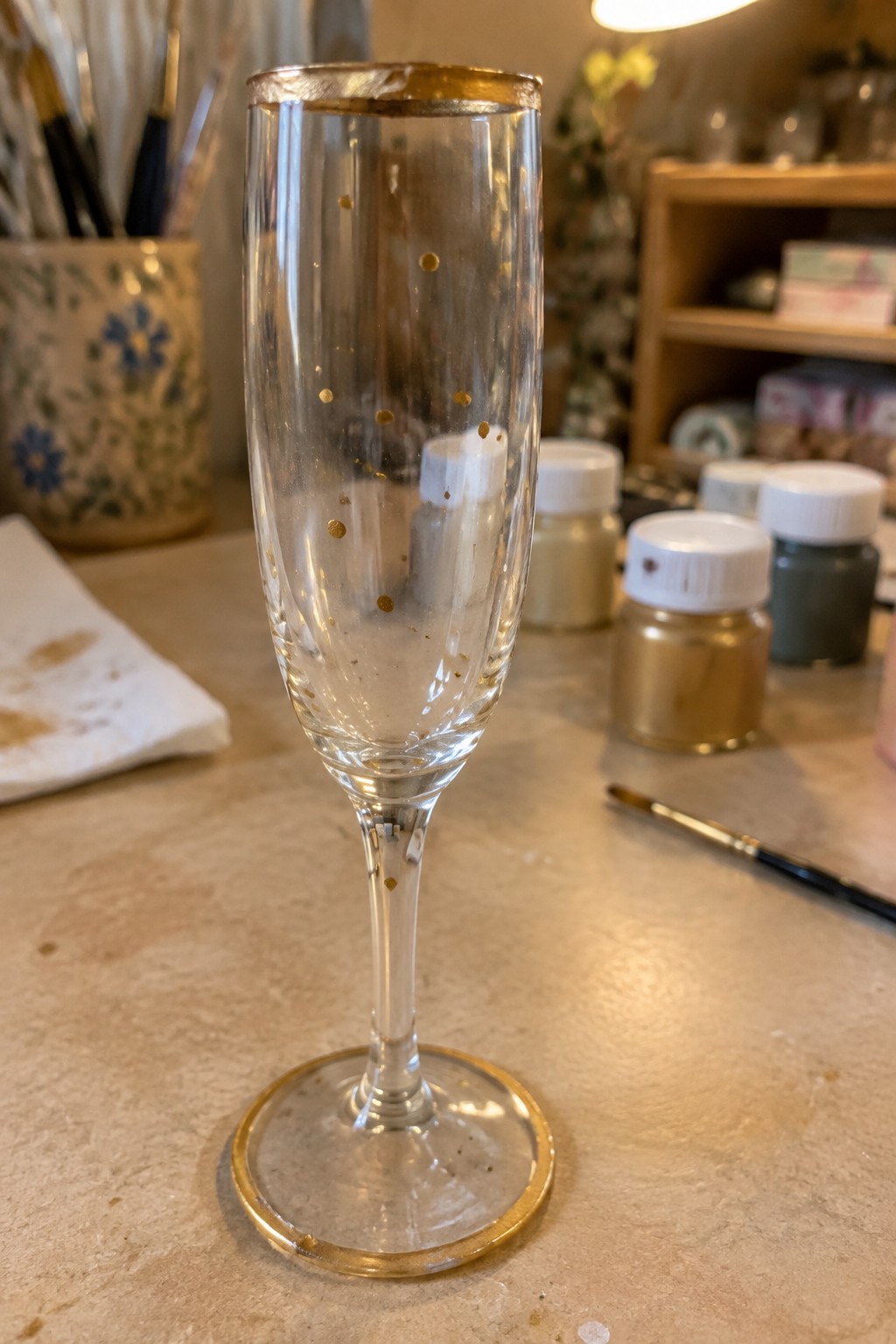 Gold-rimmed champagne flute with scattered gold dots beside paint jars and brush.