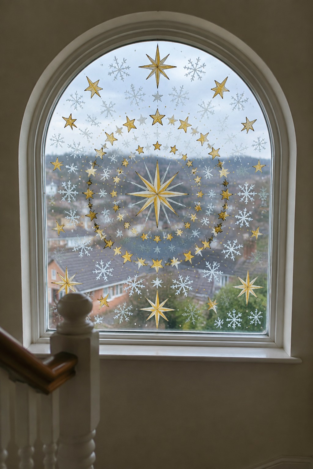 Arched window with gold and white painted stars and snowflakes in concentric circles around a large central star, seen from inside with rooftops visible beyond.