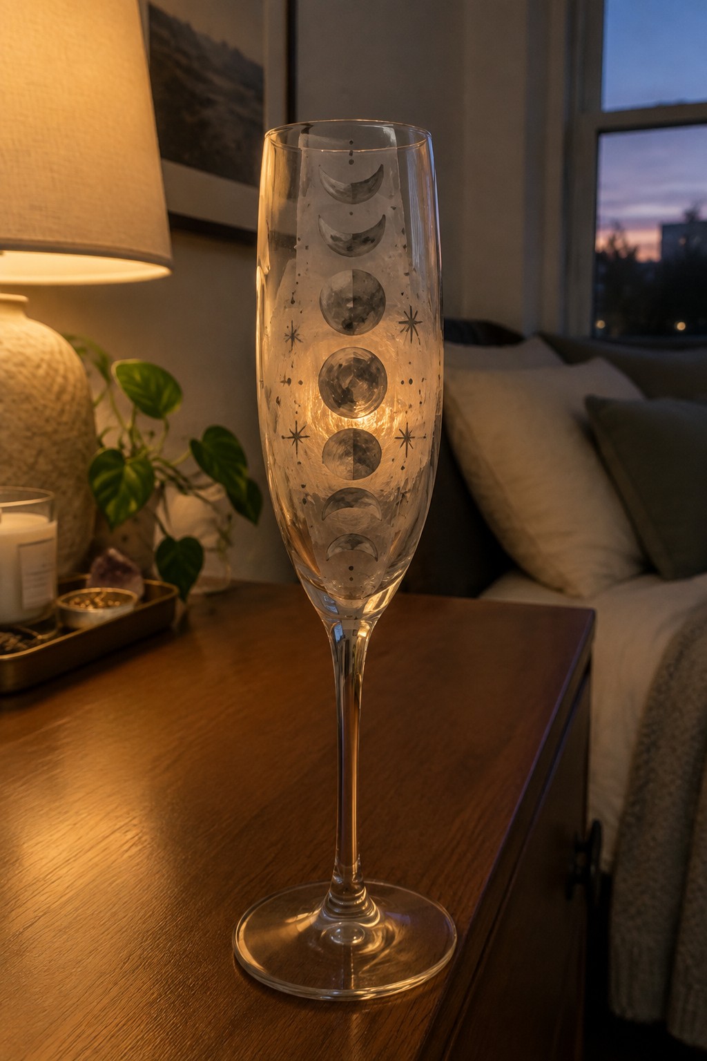 Tall champagne flute etched with moon phases glowing on wooden nightstand near lamp